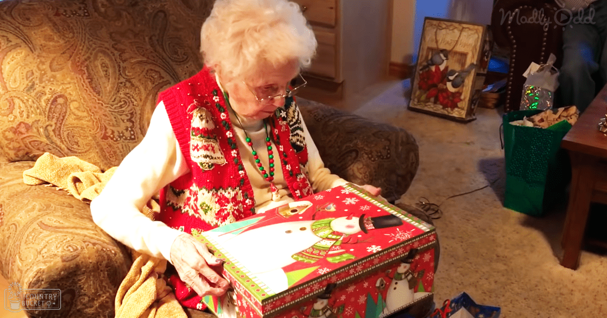 An older adult female in a festive living room setting, wearing a red and white Christmas-themed sweater vest with snowflake patterns, paired with a white shirt and beaded necklace.