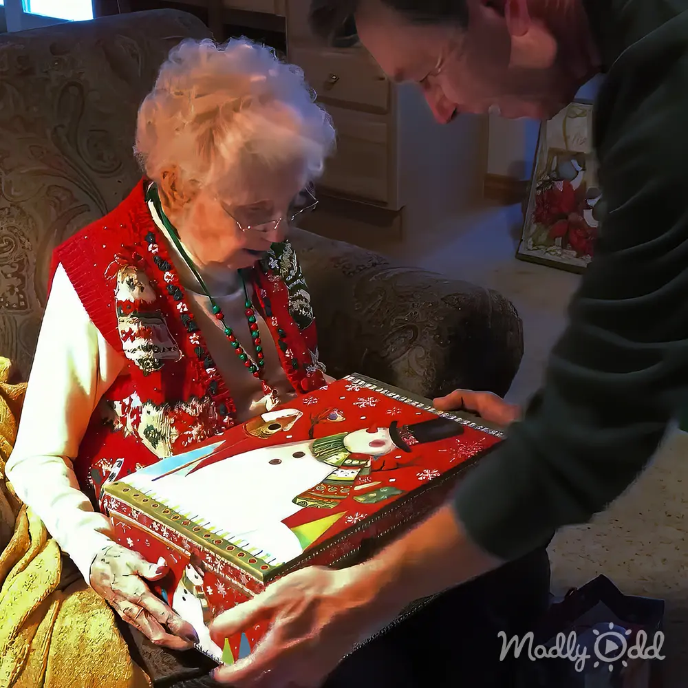 An older adult woman wearing a red Christmas-themed dress with white sleeves and festive patterns, accessorized with a green beaded necklace and glasses, in a warmly lit living room decorated for the holidays.