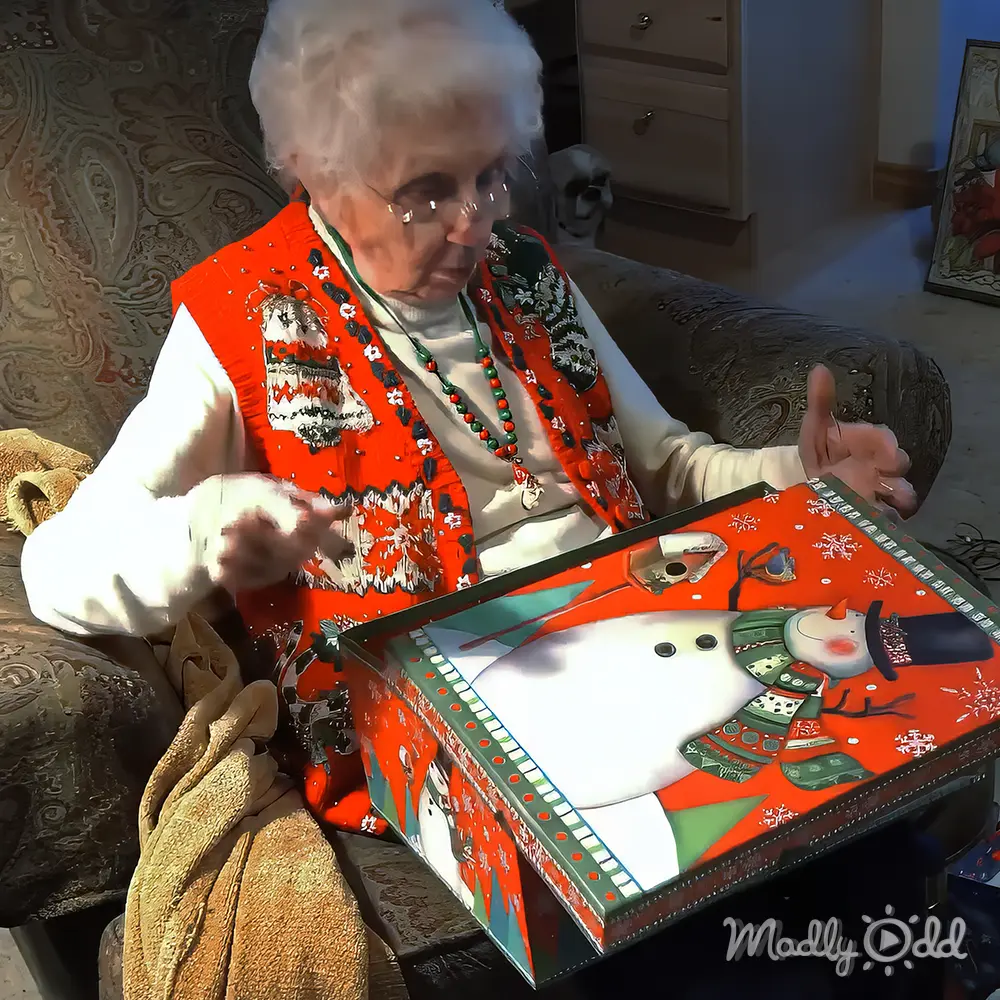 An older adult female in a living room, wearing an orange vest with festive patterns and green beads around her neck. The primary subject is surrounded by holiday-themed decor including a Christmas box.