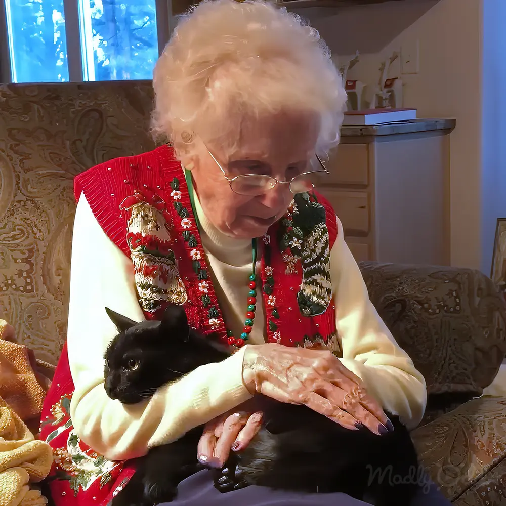 An older adult female in a living room with a red floral vest and green necklace. A black cat sits nearby on a patterned couch.