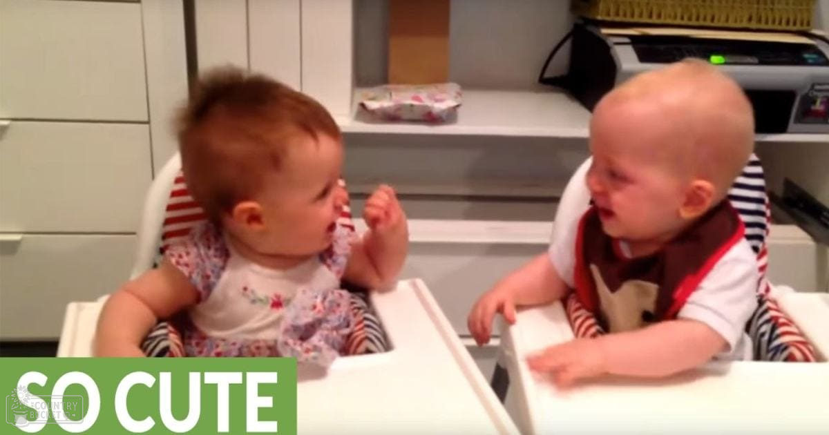 Two infants wearing white shirts with patterns and accents, sitting on white high chairs in a kitchen setting during daytime or early evening light.
