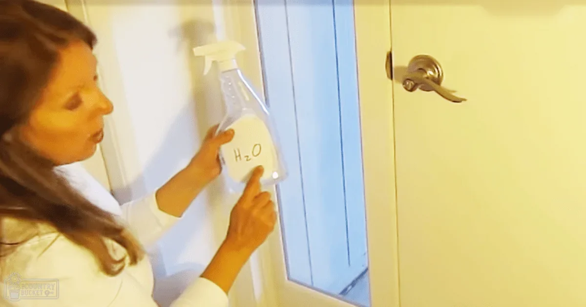 A young adult female in a white lab coat holds a clear 'H2O' spray bottle indoors, possibly in an educational or laboratory setting with artificial lighting.