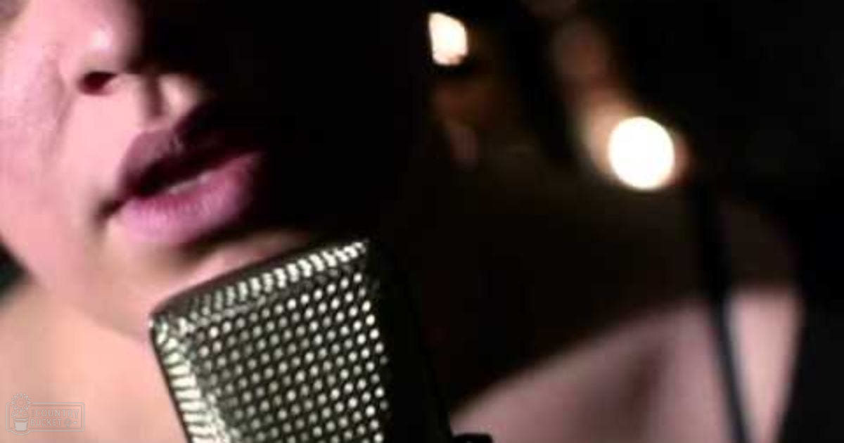 Adult female singer wearing a dark scoop-neck top in a recording studio with visible microphone and dimly lit background.