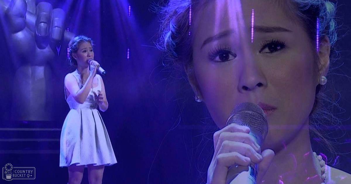 A young adult female singer wearing a white sleeveless dress with a flared skirt, performing on stage under dramatic blue and purple lighting in an indoor concert setting.