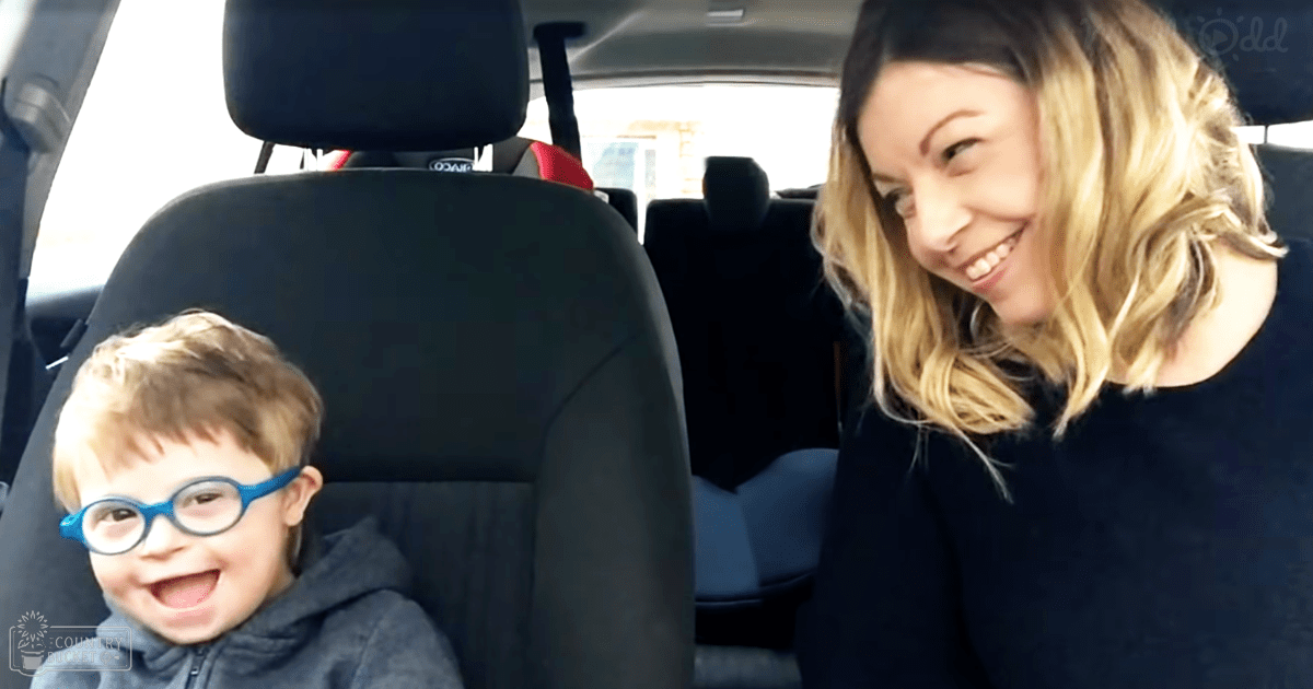 A toddler male in a dark hoodie wearing blue glasses sits inside a vehicle, accompanied by an adult female with light brown hair dressed in a dark top or dress during daytime.