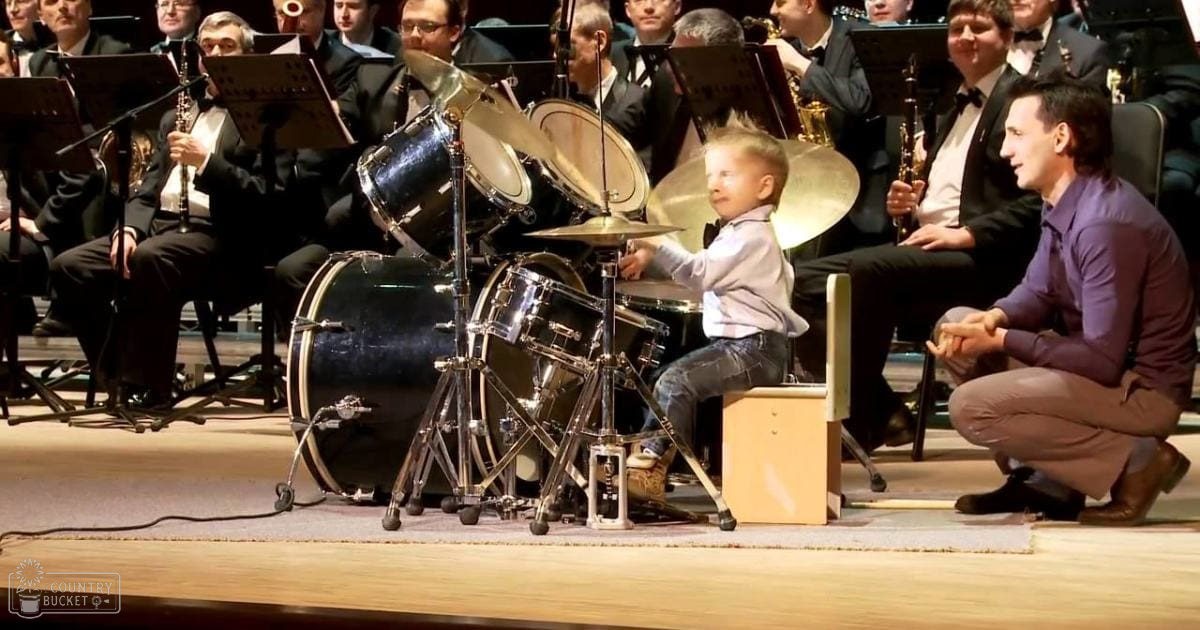 A young child plays drums in front of an orchestra at what appears to be a concert hall. The scene is filled with adult musicians seated and playing various instruments.
