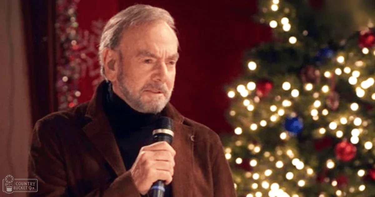 An older adult male holding a microphone, wearing a brown jacket and black turtleneck sweater in front of a decorated Christmas tree indoors during the evening holiday season.