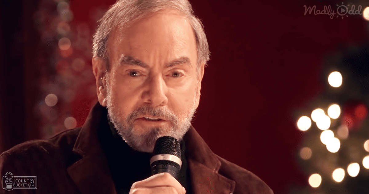 A middle-aged adult male wearing a dark, relaxed-fit jacket with a high collar in an indoor setting decorated for the holidays, holding a microphone.