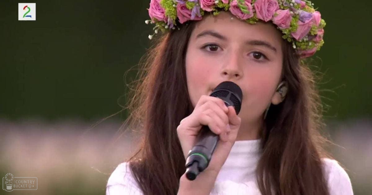 A young adult female wearing a white T-shirt with a floral crown made of pink roses and greenery, singing outdoors on what seems to be a sunny day.