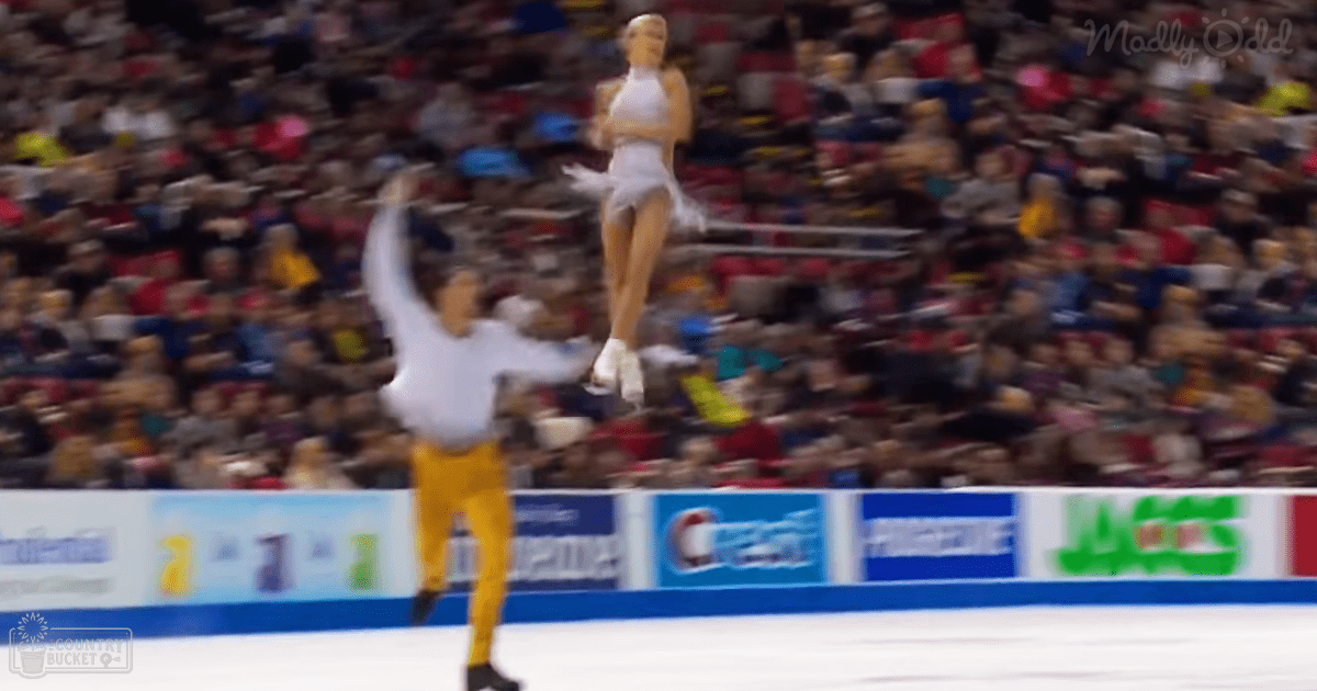 An adult male figure skater performing a lift with an adult female skater in a white outfit, set against the backdrop of an indoor arena filled with spectators under artificial light.