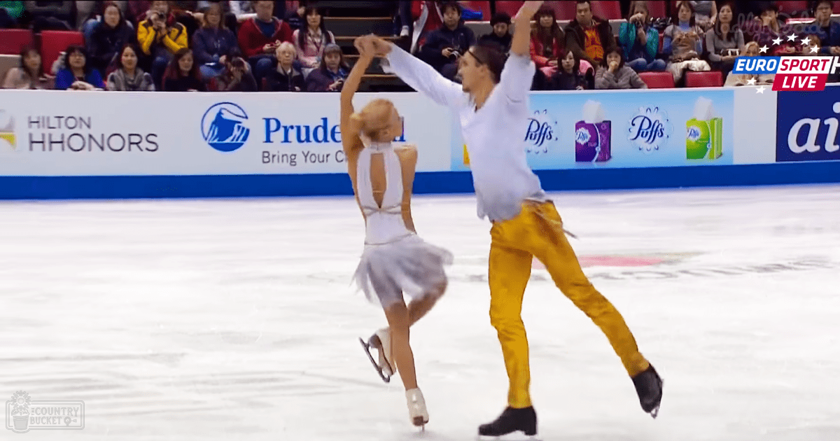 A figure skating pair in an indoor ice rink; female skater wears a white sleeveless leotard, male skater in yellow pants and a light shirt; spectators seated on red and blue seating with advertisements visible along the boards.