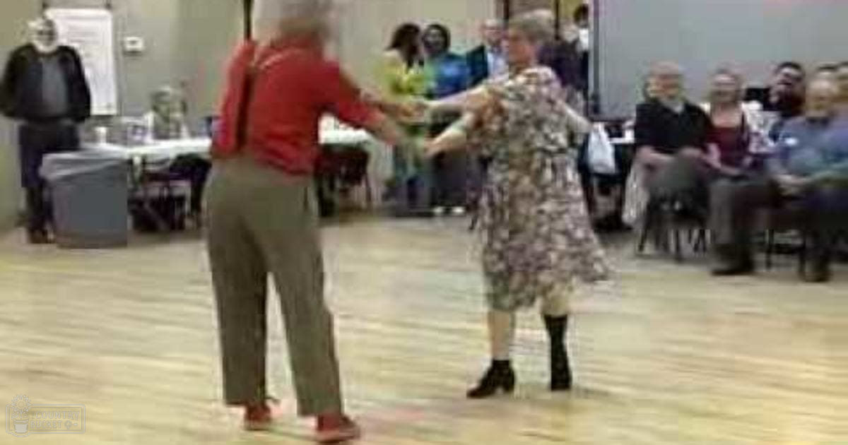 A dance scene set indoors with tables and chairs arranged in rows, suggesting a community center or event space; the lighting is bright, indicating daytime, possibly spring or summer.
