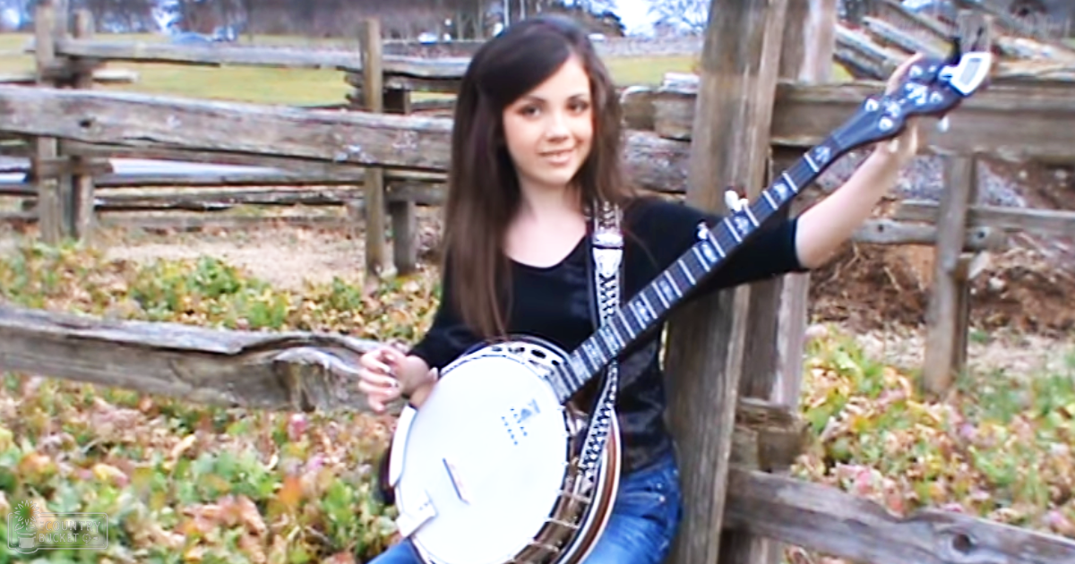 A young adult female holding a banjo outdoors against an old wooden fence, surrounded by fallen leaves in autumn or early spring under overcast daylight.
