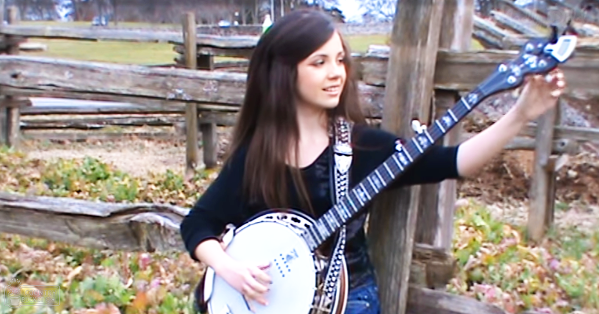 A young adult female plays a banjo in an outdoor setting with fallen leaves, suggesting autumn or early spring.