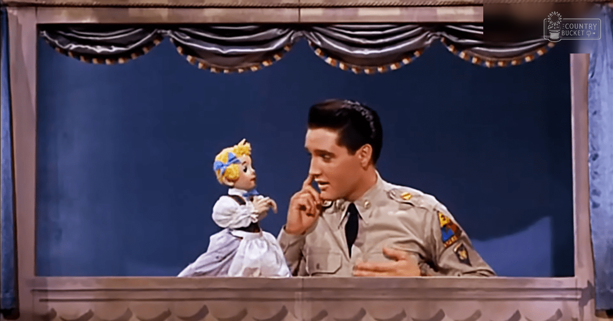 A man in a mid-20th century military-style uniform stands beside a toy doll dressed in white with blue accents, set against a simple dark blue curtain backdrop.
