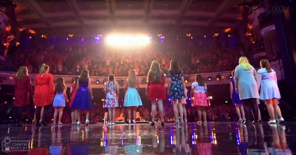 A group of young female performers in floral-patterned dresses and skirts on stage in an indoor arena or theater setting.
