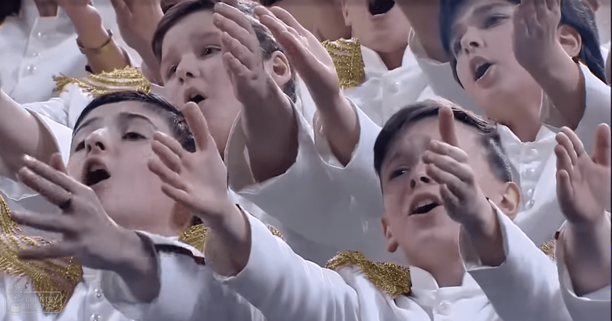Group of children in a performance setting, wearing white button-up shirts with gold embellishments at an indoor venue under artificial lighting.