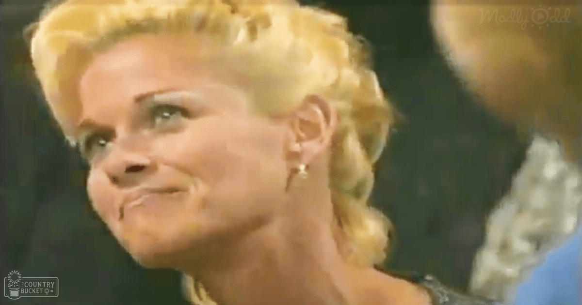 A middle-aged adult female in a blue blouse with short sleeves, wearing gold hoop earrings, stands indoors likely at an event or performance.