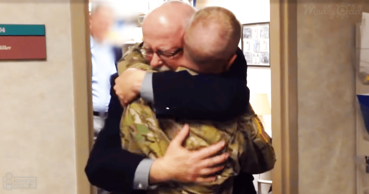 An older adult male in a dark suit jacket embraces another older adult male wearing a camouflage military uniform, set within the interior of an office or institutional environment.