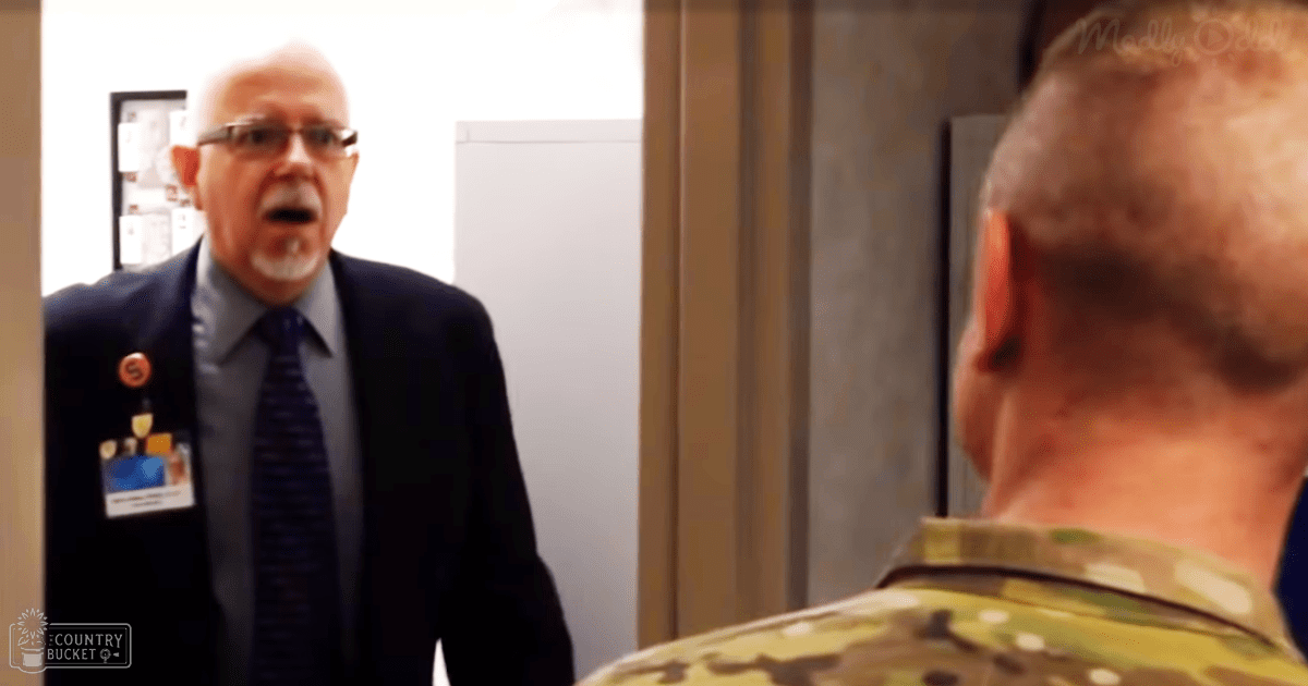 A middle-aged man in glasses wearing a dark suit jacket and patterned tie interacts with an older adult male in camouflage military uniform in an indoor office setting under daytime lighting.