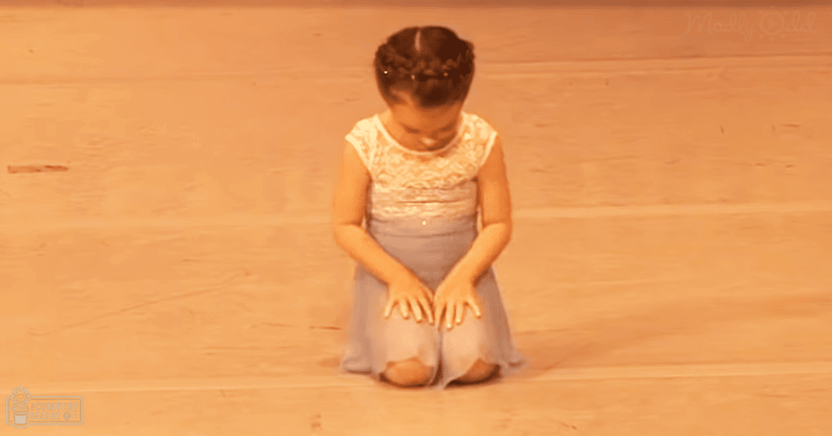 A toddler girl in a dance studio, wearing a lace-trimmed sleeveless dress and ballet slippers; light-colored attire contrasts with warm wooden floor tones.