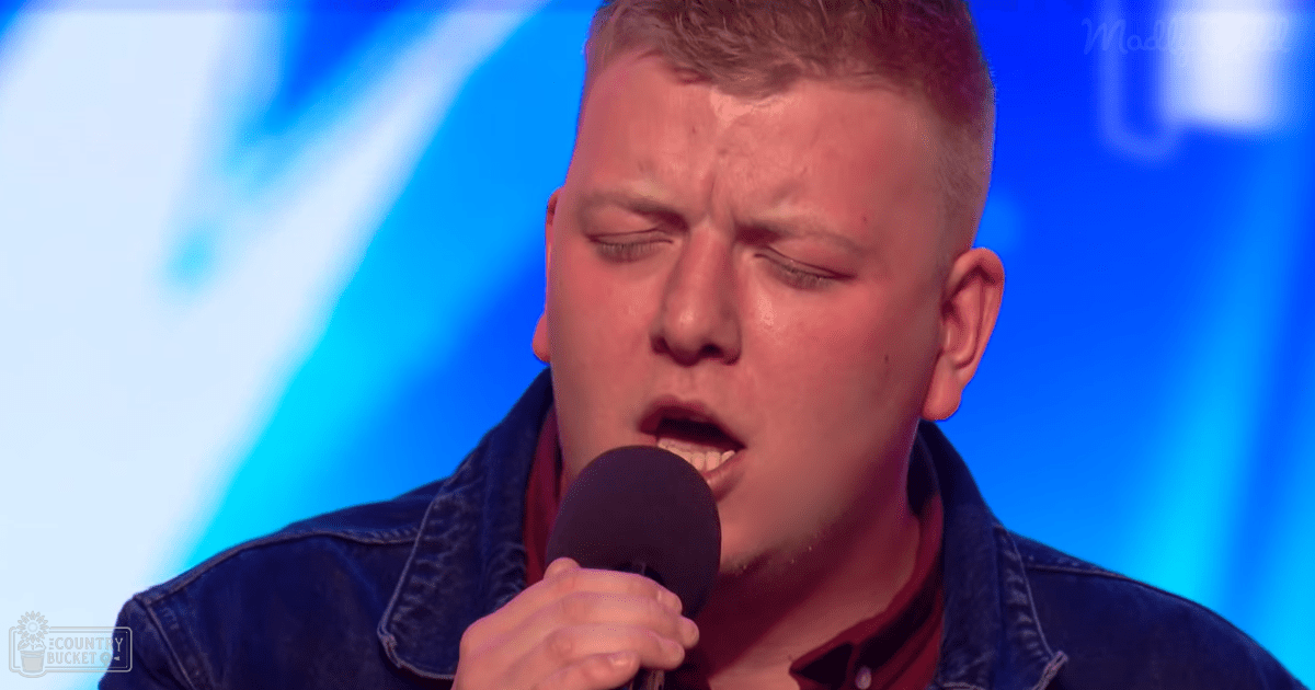 A young adult male wearing a denim jacket and red shirt sings into a microphone on what appears to be a talent show stage with blue stage lighting.