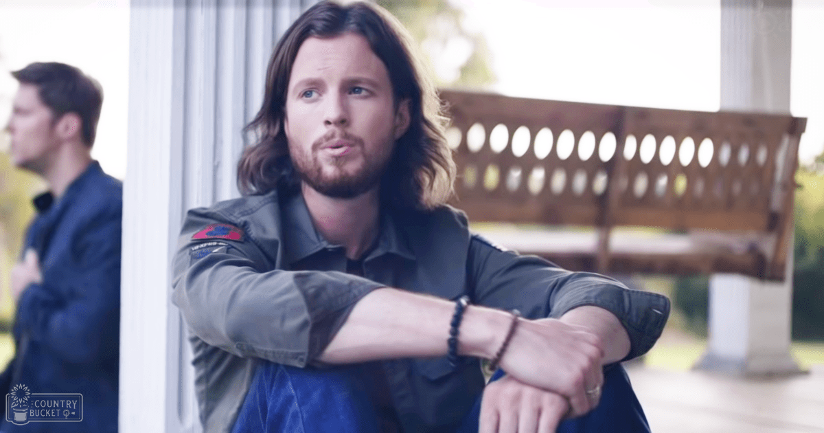 Adult male seated outdoors on a wooden bench, wearing a dark green jacket with rolled-up sleeves and blue jeans. Another adult male in a similar setting is also present, dressed in a dark blue jacket and dark pants.