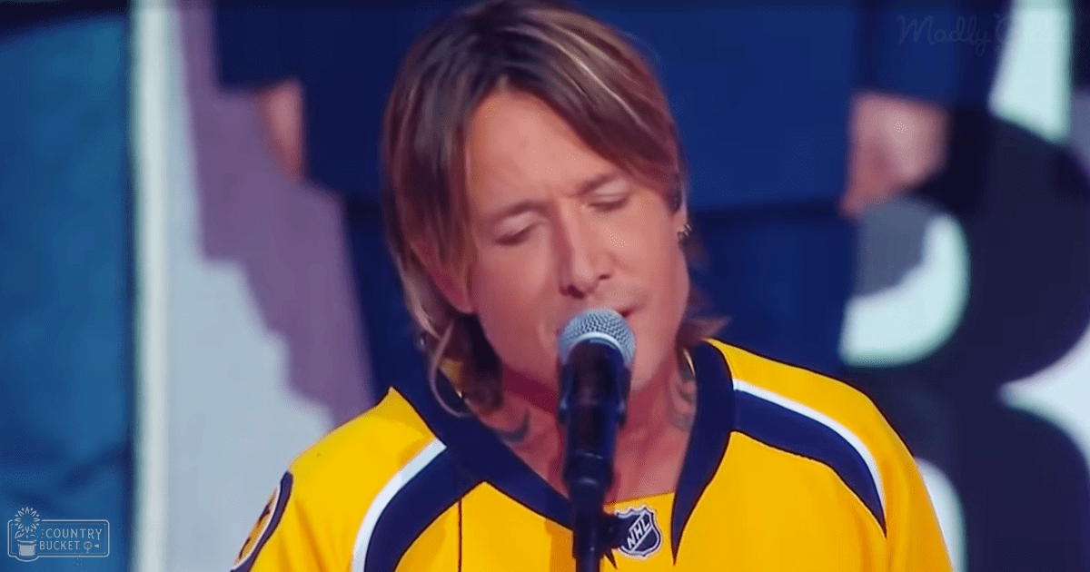 A young adult male singer in a yellow sports jersey with an NHL logo sings into a microphone at what appears to be a sports-related event. The setting is indoors, likely at a hockey arena, with stage lighting and blurred silhouettes of people in the background.