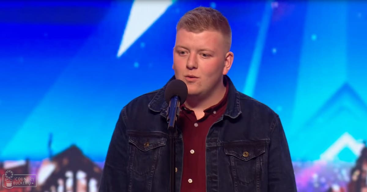 A young adult male in a dark denim jacket and maroon shirt on stage, possibly performing or speaking.