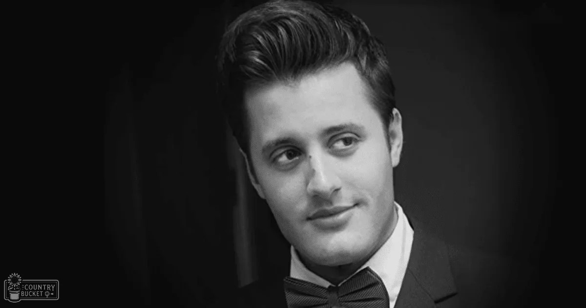 A young adult male in a dark suit jacket, white dress shirt, black bow tie, indoors with controlled lighting.