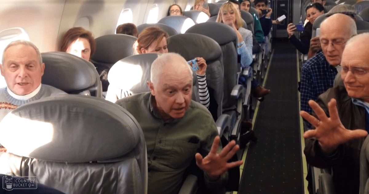 An older adult male in a green button-up shirt seated inside an airplane cabin with passengers around him, during daytime travel.