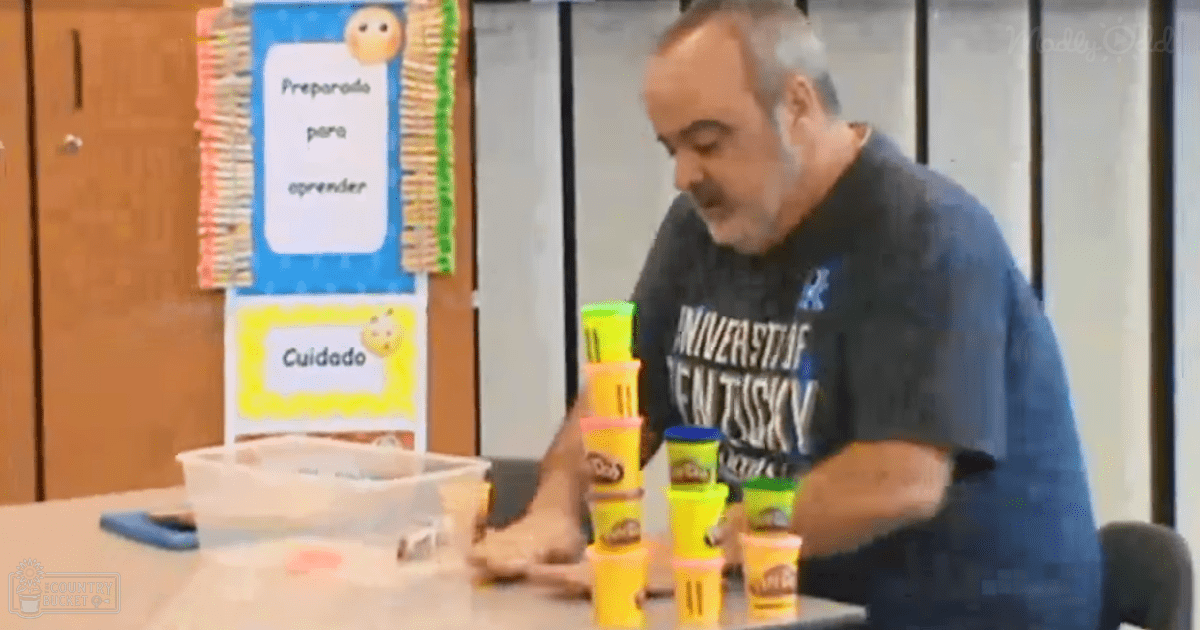 An older adult male wearing a dark blue 'University of Kentucky' shirt in a classroom setting with educational posters and decorations, including phrases like 'Preparado para aprender'.