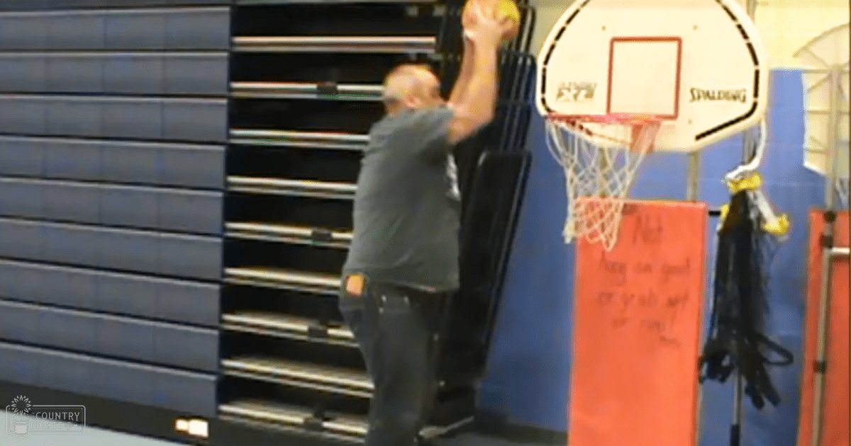 A middle-aged male wearing a gray casual-style t-shirt plays basketball in an indoor gymnasium with blue wall panels, a Spalding basketball hoop, and a red padded mat.