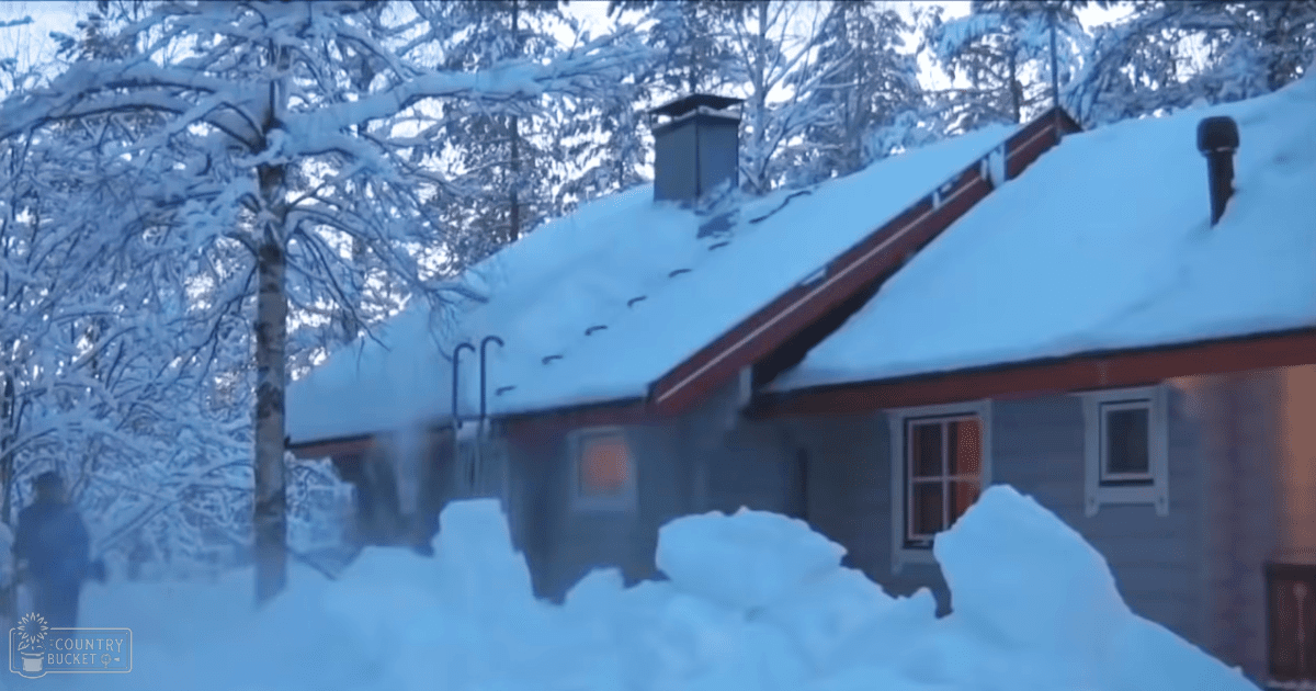 A snow-covered cabin with a chimney, emitting light through its windows in a winter landscape surrounded by snowdrifts and snow-capped trees.