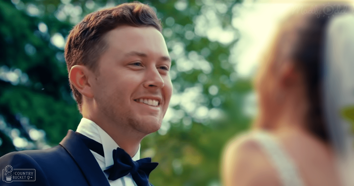 A young adult male in a dark blue tuxedo at an outdoor event with greenery and lush trees in the background.