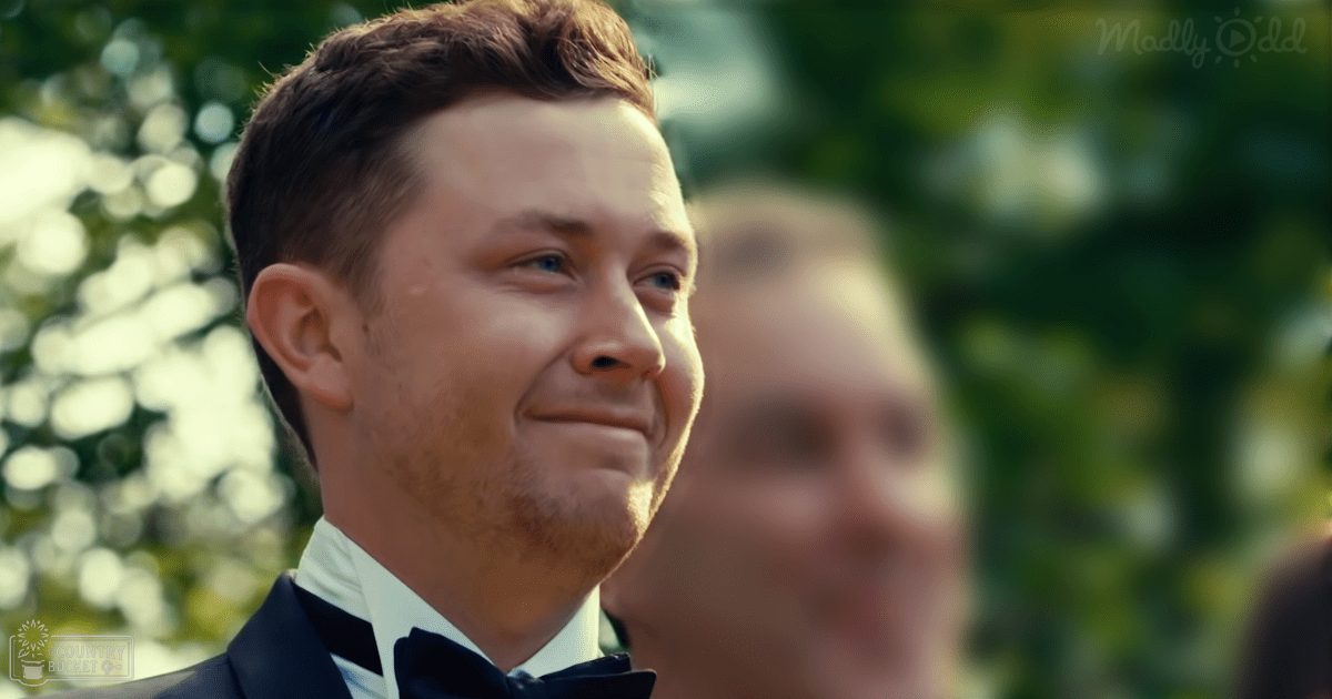 A young adult male in a dark suit with a white shirt and black bow tie stands outdoors amidst greenery, wearing glasses.