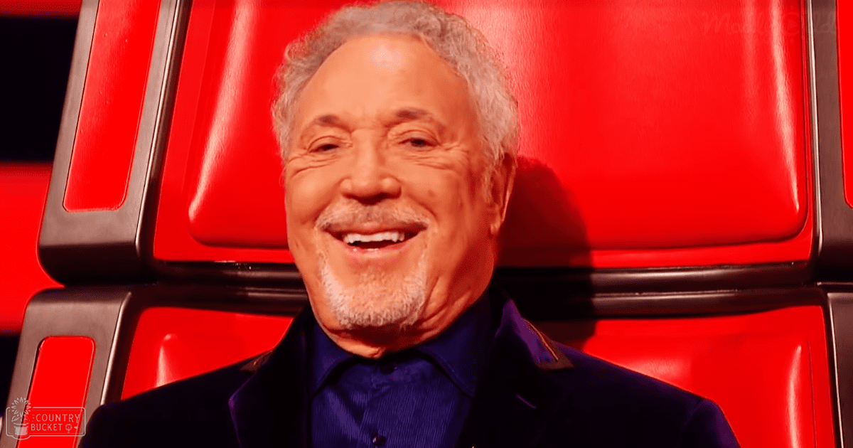 An older adult male in a modern formal attire, seated on stage or in a judging panel area within an indoor studio environment typical of talent shows or reality TV programs.