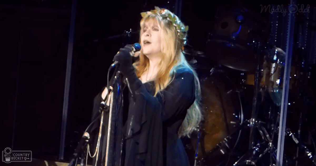A middle-aged female performer sings on stage, wearing a long-sleeved black dress with full-length sleeves and a high neckline, adorned with a floral headband and black gloves.