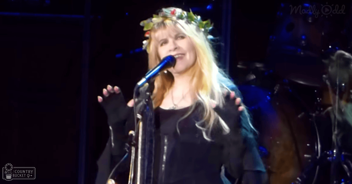A middle-aged female performer wearing a black leather jacket, fingerless gloves, and a floral crown, singing into a microphone at an indoor concert with stage lighting.