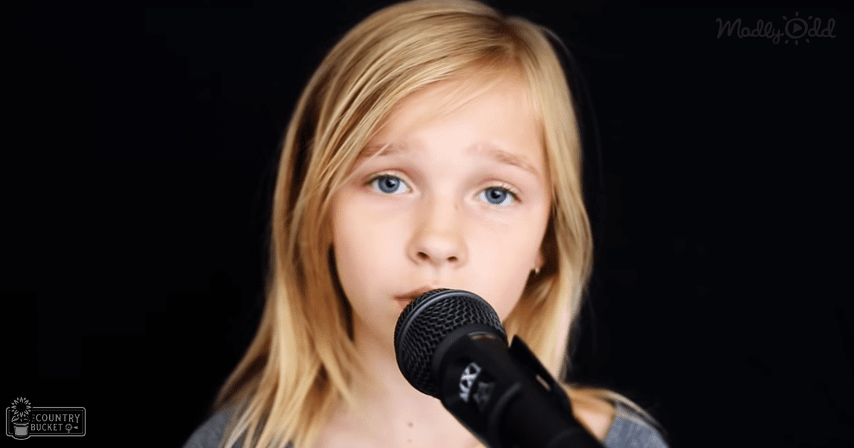 A child in a relaxed, long-sleeved top of grayish-blue color with subtle patterns stands indoors against a dark background under focused artificial lighting.
