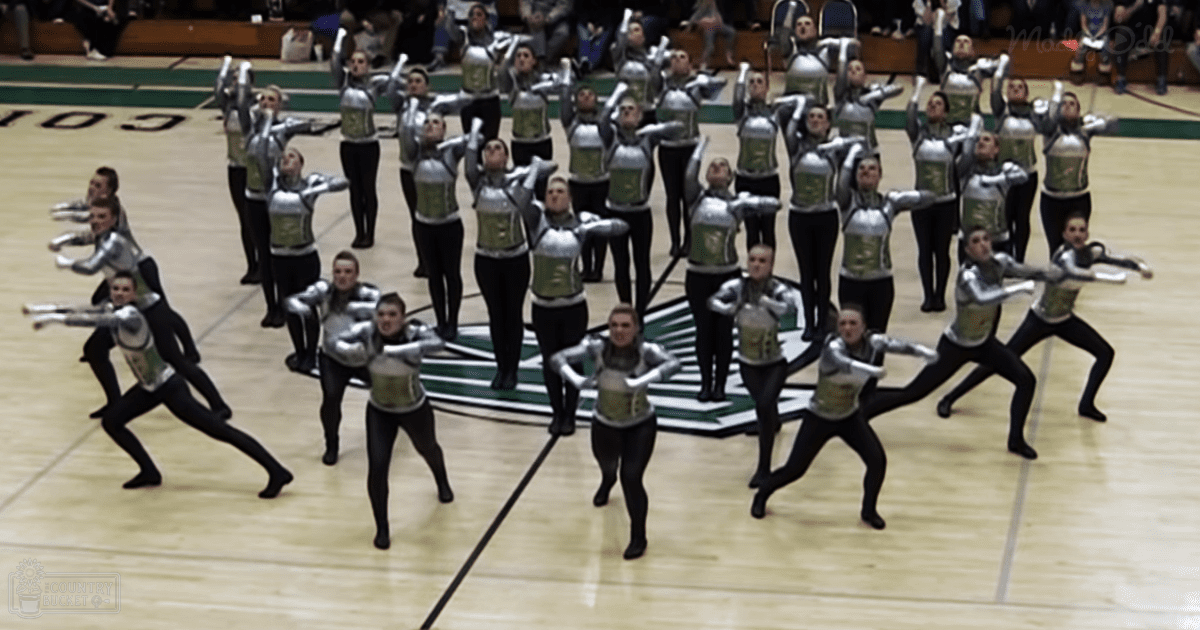 Young adult female dancers performing in a gymnasium, wearing matching uniforms of black leggings, silver and green tops with white trim, and accessories that match the uniform colors.