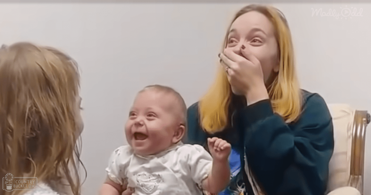 An infant wearing a white Superman onesie indoors, accompanied by two adults with long blonde hair, surrounded by formal decor and light-colored walls.