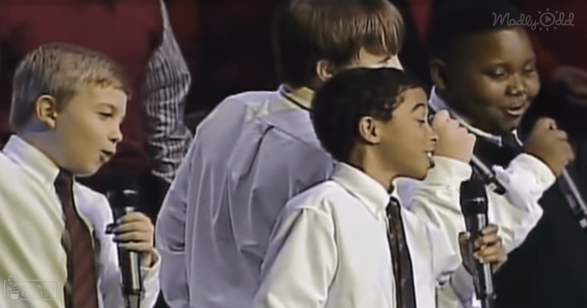 A group of children, ages 8-10 years old, dressed in formal attire with white shirts and dark ties, are singing into microphones at an indoor performance venue. The setting is focused on the children, with even artificial lighting and blurred background.