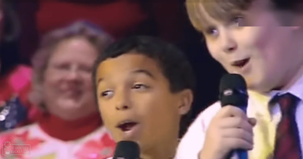 A young boy wearing a patterned white shirt sings into a microphone in what appears to be a performance setting with warm indoor lighting, while an adult female and another child are also present.