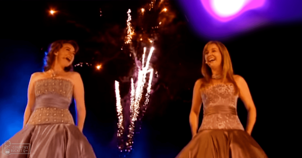 Two young adult female models dressed in elegant floor-length gowns, celebrating at an outdoor night event with fireworks and purple lighting.