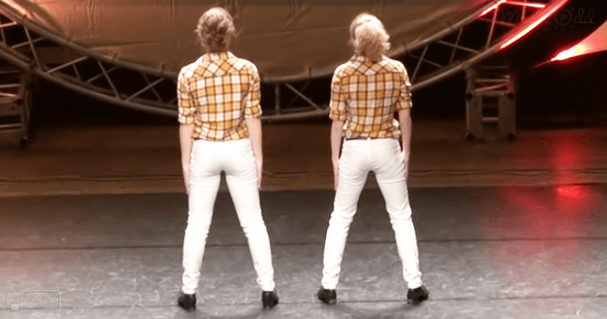 Two young adult female performers in a casual, checkered shirt ensemble with slim-fit pants and black slip-on shoes on a stage with metal scaffolding.
