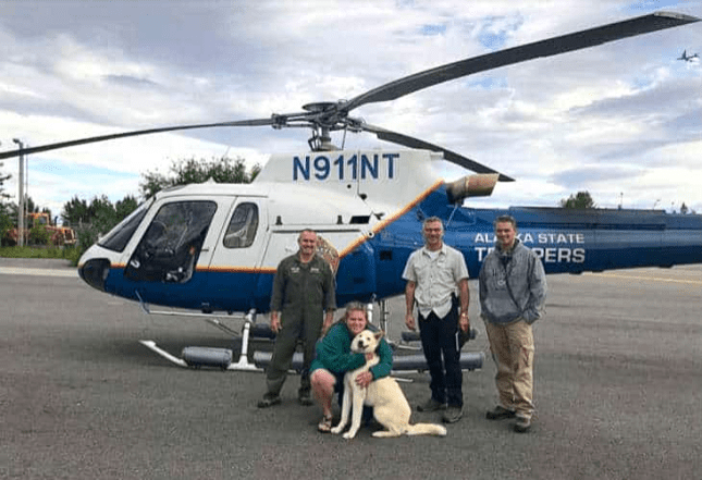 A golden retriever mix dog, likely an adult, stands in front of a helicopter marked 'N911NT' and 'Alaska State Troopers,' on an airfield with partly cloudy skies.