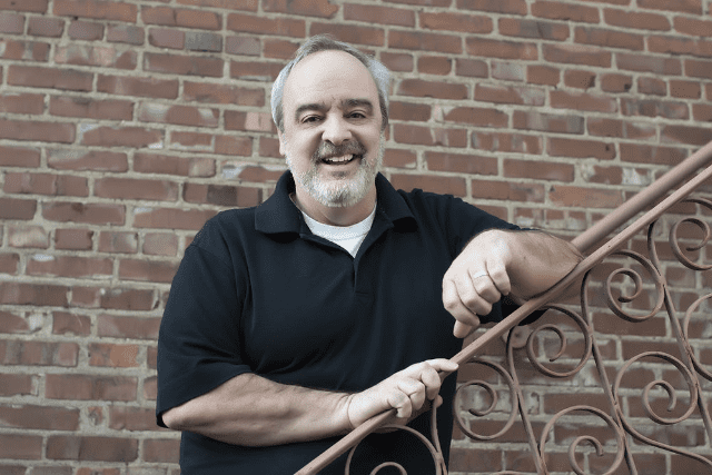 Middle-aged adult male holding an ornate metal railing outdoors against a brick wall in modern casual wear.