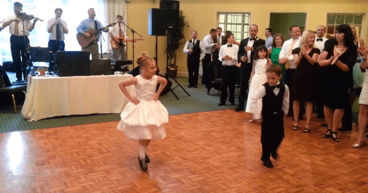 A young girl in a white dress dances at an indoor wedding reception with musicians and guests in formal attire, under natural daylight.