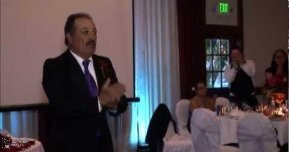 A middle-aged man in a dark suit and purple tie giving a speech at an indoor wedding reception with formal decor, surrounded by other attendees seated around white-clothed tables adorned with floral centerpieces.